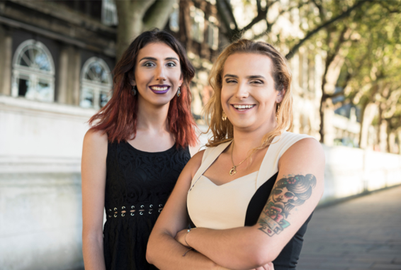 Two trans people look smiling at the camera, experiencing a quiet joy.