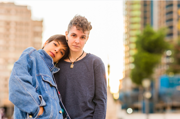 Two people stand in the foreground of the picture, the city out of focus in the background. One person has a short haircut and wears a blue jean jacket, leading back on the shoulder of a person with short quaffed curly hair and wears a grey long sleeve shirt.