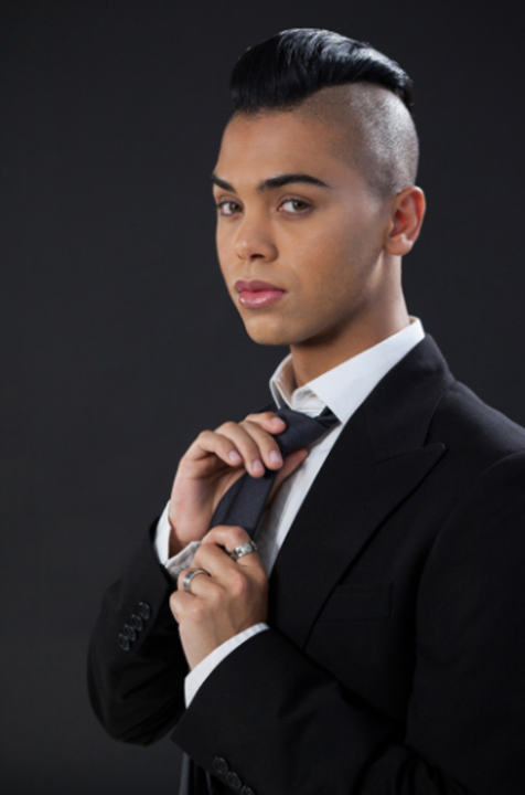 This masculine Latino person wears a black suit that fades into the black background. He adjusts his slim black tie and gazes casually into the camera. 