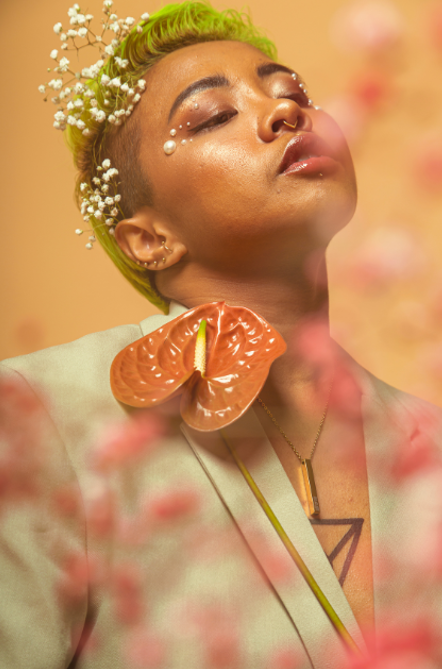 In this close-shot, a person looks up with a relaxed expression and closed eyes at a soft light. They have a large red laceleaf flower on their blazer and small white baby breath flowers in their short green hair. They are calm and serene.