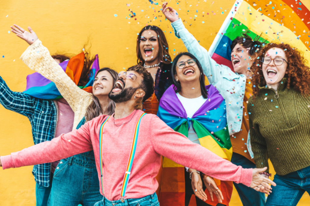 Seven people stand tightly together. Dressed in colorful clothing and waving pride flags or using pride flags as capes, the people radiate queer joy. They are smiling with open mouths and have outstretched arms. 