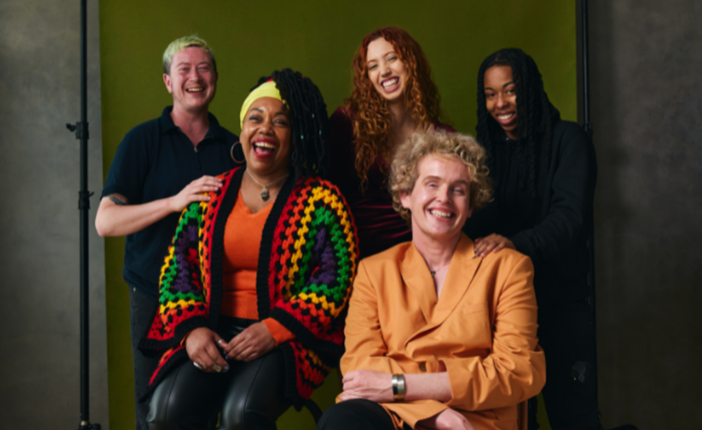 In this group photo, three people stand behind two seated people, all smiling or laughing. There are people of many gender expressions represented. 