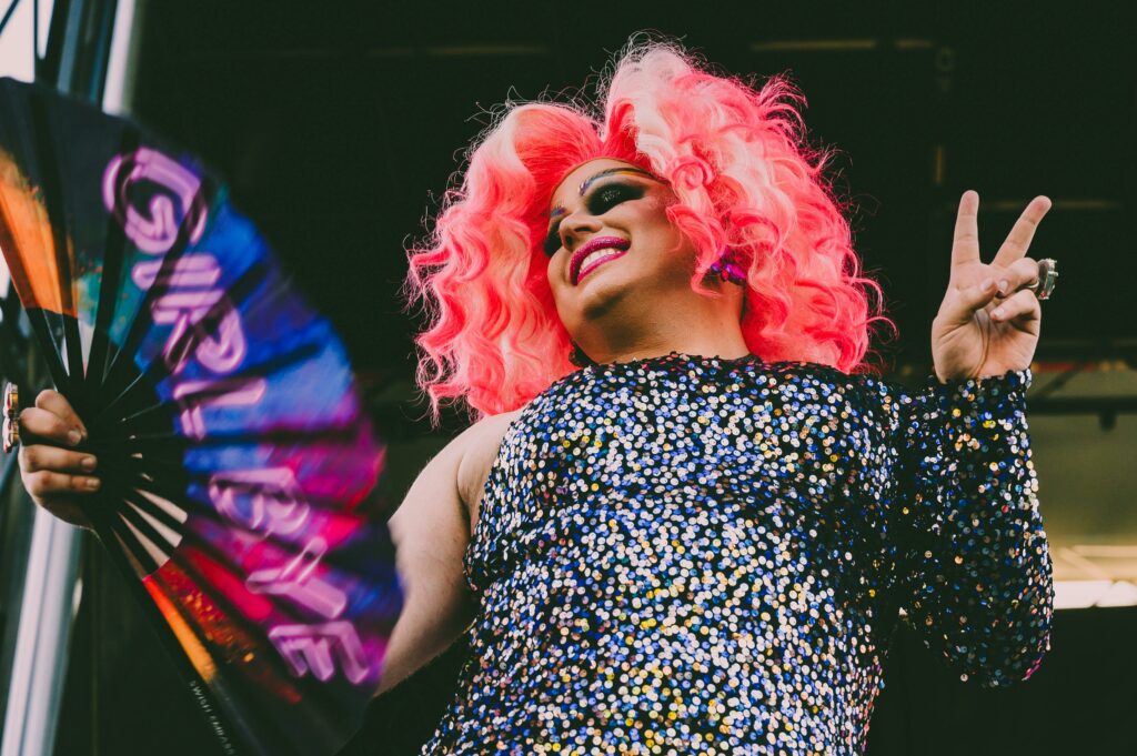 Drag queen with a sparkling top and bright pink wig holding a hand fan.